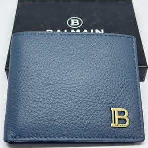 Może przedstawiać: Niebieski skórzany portfel ze złotym logo "B" i nazwą marki "Balmain". Portfel jest prezentowany przed czarnym pudełkiem z nazwą marki i logo. Portfel ma teksturowaną powierzchnię.