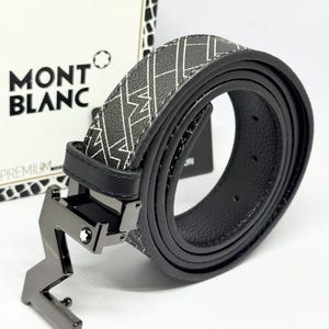 May include: A black belt with a repeating white geometric pattern and a black metal buckle shaped like the letter M. The buckle features a small white star emblem. The belt is coiled in front of a white box labeled MONT BLANC PREMIUM.