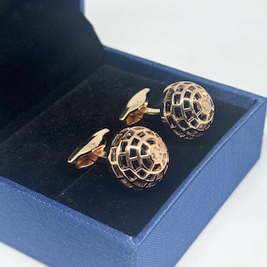 Può includere: Un paio di gemelli color oro rosa con un design sferico a griglia e un piccolo emblema a forma di stella sulla parte superiore. I gemelli sono presentati in una scatola blu con un rivestimento interno in velluto nero.