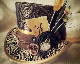 May include: A black and gold top hat with a paisley pattern. The hat is decorated with a brown ribbon, a small clock, a black rose, and a brown paper tag with the text "1/6".