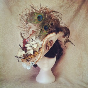 May include: A decorative hat featuring a miniature sailing ship, peacock feathers, and a Union Jack flag. The hat has a cream-colored brim with a map design and a brown ribbon. The hat is displayed on a white mannequin head.
