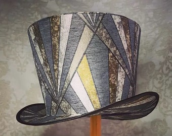 May include: A tall, gray top hat with a geometric pattern in shades of brown and gold. The hat is displayed on a wooden stand.
