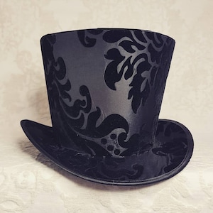 May include: A black top hat with a damask pattern. The hat is made of a smooth, silky fabric.