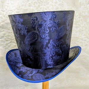 May include: A tall top hat with a paisley pattern in shades of blue and black. The hat has a wide brim with a blue trim. The hat is made of a fabric with a detailed, swirling design.