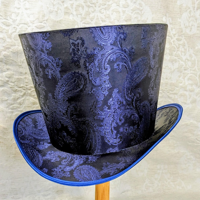 May include: A tall top hat with a paisley pattern in shades of blue and black. The hat has a wide brim with a blue trim. The hat is made of a fabric with a detailed, swirling design.