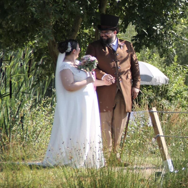 May include: A bride in a white wedding dress with a veil and a bouquet of flowers, and a groom in a brown suit and top hat, walking together outdoors. The couple is holding hands and smiling. The background includes greenery and a small bridge.