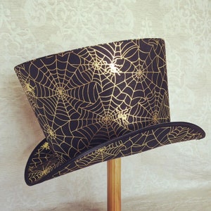 May include: A black top hat with a gold spiderweb pattern. The hat has a wide brim and a tall crown.