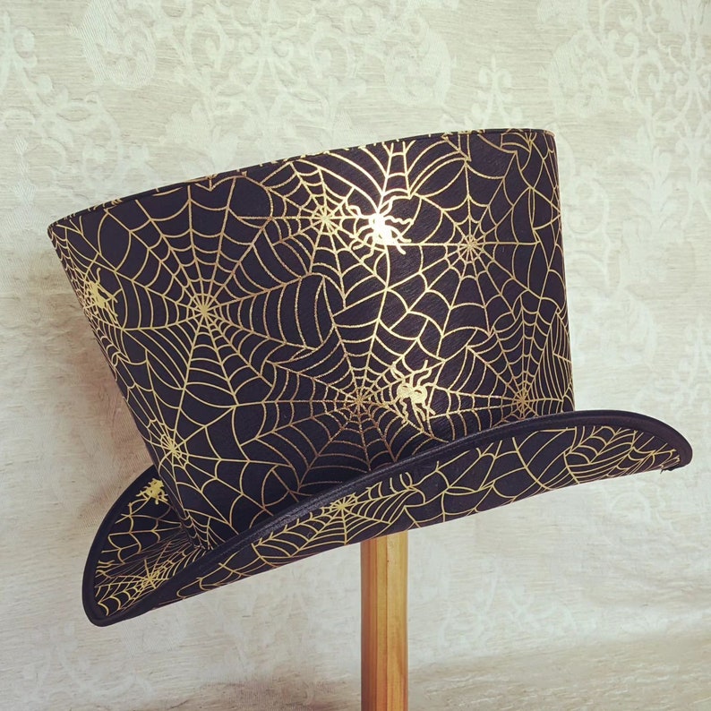 May include: A black top hat with a gold spiderweb pattern. The hat has a wide brim and a tall crown.
