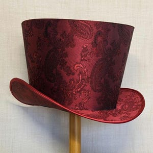 K&ouml;nnte beinhalten: Ein bordeauxfarbener Zylinderhut mit Paisley-Muster. Der Hut hat eine breite Krempe und eine hohe, zylindrische Krone. Der Hut ist aus gl&auml;nzendem Stoff gefertigt und wird auf einem h&ouml;lzernen Hutst&auml;nder pr&auml;sentiert.