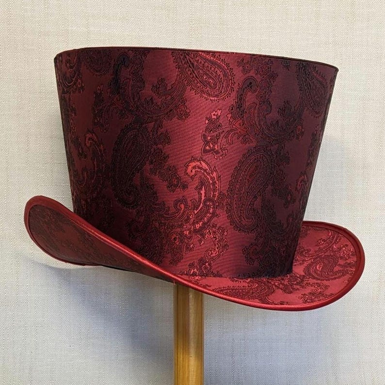 K&ouml;nnte beinhalten: Ein bordeauxfarbener Zylinderhut mit Paisley-Muster. Der Hut hat eine breite Krempe und eine hohe, zylindrische Krone. Der Hut ist aus gl&auml;nzendem Stoff gefertigt und wird auf einem h&ouml;lzernen Hutst&auml;nder pr&auml;sentiert.