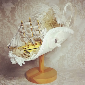 K&ouml;nnte beinhalten: Ein skurriler wei&szlig;er Hut mit einem Miniatur-Holzsegelschiff, verziert mit Spitze, einer Schleife und einem dekorativen Knopf. Der Hut ist mit wei&szlig;en Federn verziert und wird auf einem Holzst&auml;nder pr&auml;sentiert, ein einzigartiges und k&uuml;nstlerisches Accessoire.