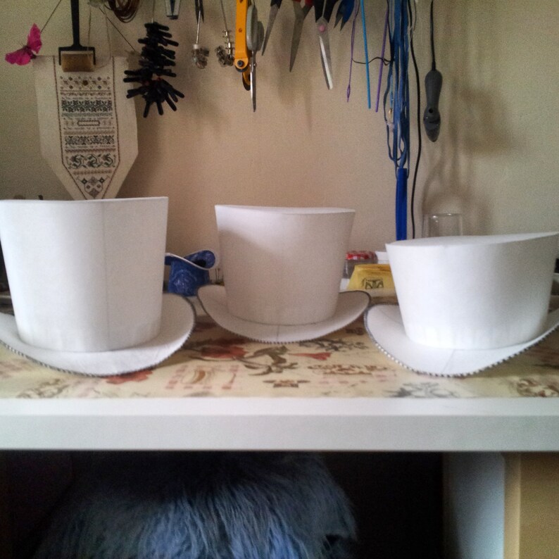 May include: Three white top hats of different sizes sitting on a table with a floral pattern tablecloth.