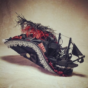 May include: A black hat with a miniature ship, red and black feathers, and white lace trim. The hat features a gold button and red ribbon accents, creating a unique, decorative accessory.