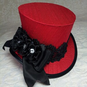 May include: A red top hat with a black brim and accents. The hat features a black ribbon, a black flower with rhinestones, and black lace trim. The hat is a decorative accessory.