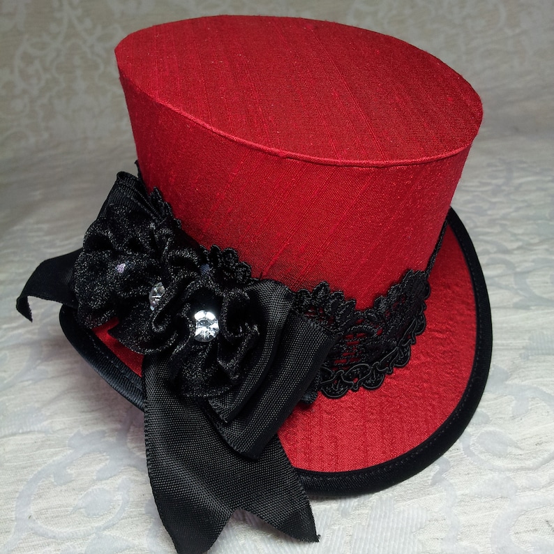 May include: A red top hat with a black brim and accents. The hat features a black ribbon, a black flower with rhinestones, and black lace trim. The hat is a decorative accessory.