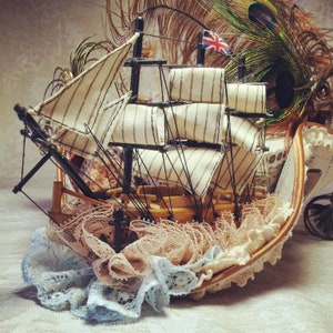 May include: A miniature wooden sailboat with cream-colored striped sails and a Union Jack flag. The boat is adorned with delicate lace in shades of peach and blue, creating a whimsical nautical theme.