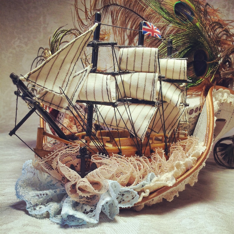 May include: A miniature wooden sailboat with cream-colored striped sails and a Union Jack flag. The boat is adorned with delicate lace in shades of peach and blue, creating a whimsical nautical theme.
