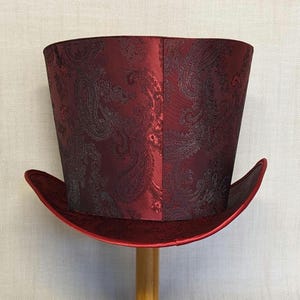 K&ouml;nnte beinhalten: Ein bordeauxfarbener Zylinderhut mit Paisley-Muster. Der Hut hat eine breite Krempe und eine hohe, zylindrische Krone. Der Hut ist aus gl&auml;nzendem Stoff gefertigt und wird auf einem Holzst&auml;nder pr&auml;sentiert.
