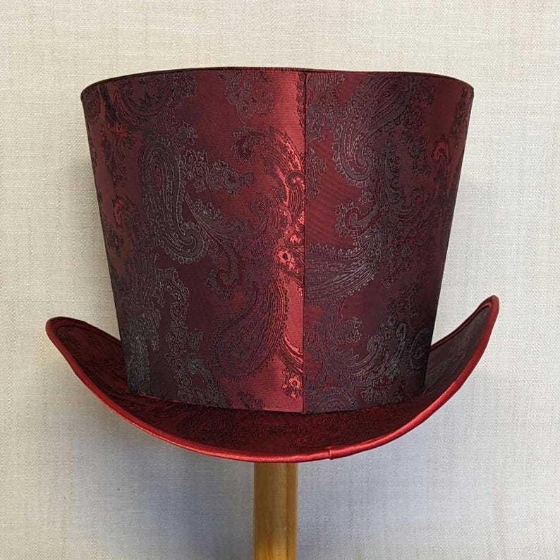 K&ouml;nnte beinhalten: Ein bordeauxfarbener Zylinderhut mit Paisley-Muster. Der Hut hat eine breite Krempe und eine hohe, zylindrische Krone. Der Hut ist aus gl&auml;nzendem Stoff gefertigt und wird auf einem Holzst&auml;nder pr&auml;sentiert.