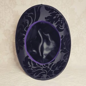 May include: A black velvet hat with a purple satin trim and a black damask pattern.