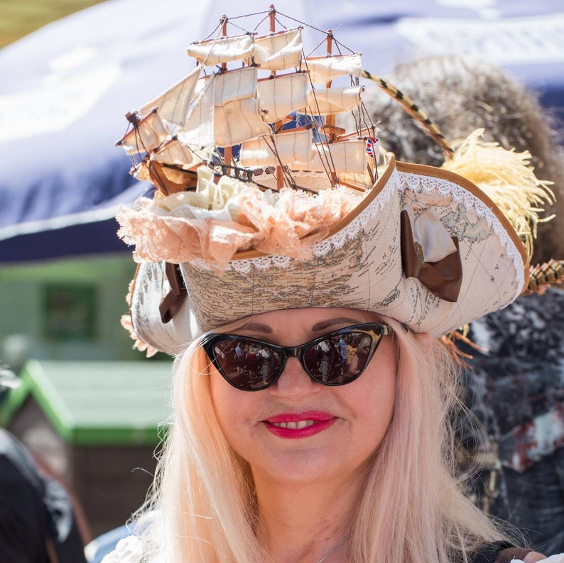 May include: A whimsical hat featuring a miniature sailing ship atop a map-printed brim. The hat is adorned with lace, ribbons, and a small British flag. The wearer is also wearing black cat-eye sunglasses and has pink hair.