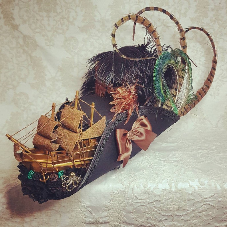 K&ouml;nnte beinhalten: Ein schwarzer Piratenhut mit einem goldenen Schiff, einer braunen Schleife und dekorativen Federn. Der Hut hat ein goldenes Schiffsmodell, eine braune Satinschleife und ist mit schwarzen, braunen und gr&uuml;nen Federn verziert, was ein skurriles und theatralisches Design ergibt.