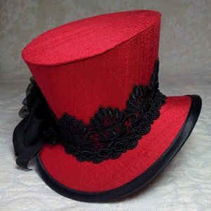 May include: A red top hat with a black lace band and a black ribbon bow. The hat has a black brim and a textured fabric. This hat is suitable for cosplay or costume events.
