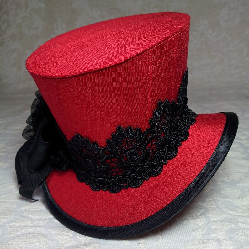 May include: A red top hat with a black lace band and a black ribbon bow. The hat has a black brim and a textured fabric. This hat is suitable for cosplay or costume events.