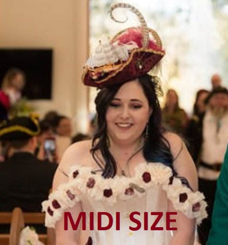 K&ouml;nnte beinhalten: Ein burgunderfarbener und goldener Piratenhut mit wei&szlig;en Schiff- und Federakzenten. Der Hut wird von einer Person getragen, die ein wei&szlig;es Kleid mit floralen Akzenten tr&auml;gt. Der Text "MIDI SIZE" ist auf dem Kleid zu sehen.