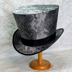 May include: A black and silver top hat with a paisley pattern. The hat is on a wooden stand.