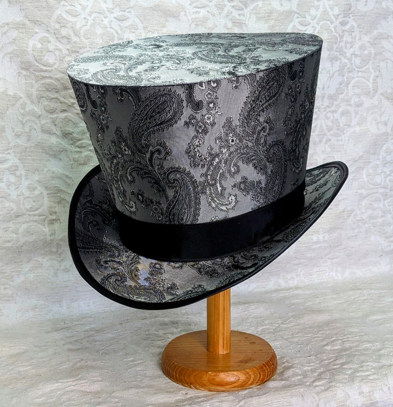 May include: A black and silver top hat with a paisley pattern. The hat is on a wooden stand.