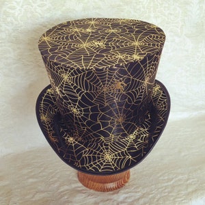 May include: A black top hat with a gold spiderweb pattern. The hat is on a wooden stand.