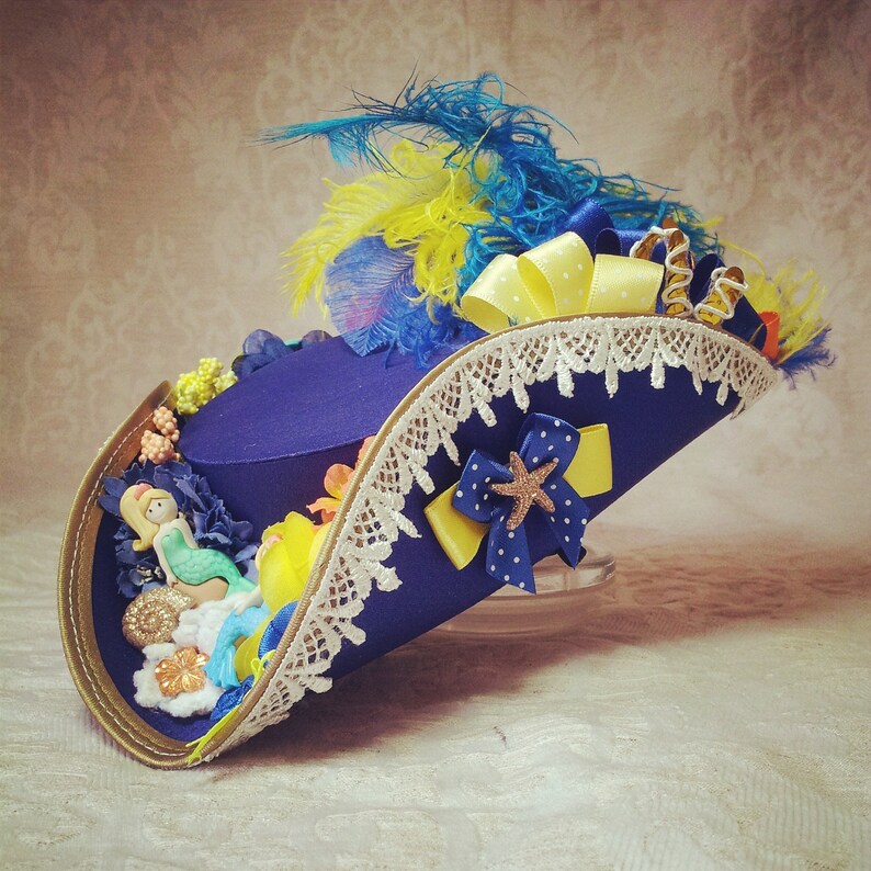 May include: A navy blue pirate hat with a gold trim, adorned with a mermaid figurine, yellow and blue feathers, and a white lace trim. The hat features a yellow polka dot bow and a blue bow with a starfish accent.