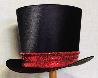 Peut inclure: Un chapeau haut de forme noir avec un large bord et une bande rouge à paillettes. Le chapeau est fait d'un matériau brillant et est présenté sur un support en bois. Ce chapeau classique est adapté aux spectacles ou aux soirées costumées.