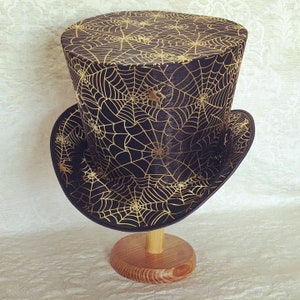 May include: A black top hat with a gold spiderweb pattern. The hat is on a wooden stand.