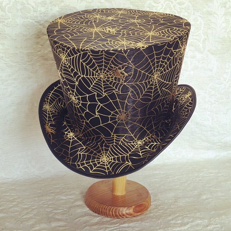 May include: A black top hat with a gold spiderweb pattern. The hat is on a wooden stand.