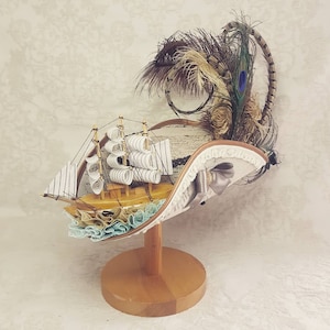 K&ouml;nnte beinhalten: Dekorativer Hut mit einem Miniatur-Holzsegelschiff mit wei&szlig;en Segeln und einer hellblau-beigen R&uuml;schenbasis. Der Hut ist mit Federn, einer Schleife und Spitzenbesatz verziert und wird auf einem Holzst&auml;nder pr&auml;sentiert.