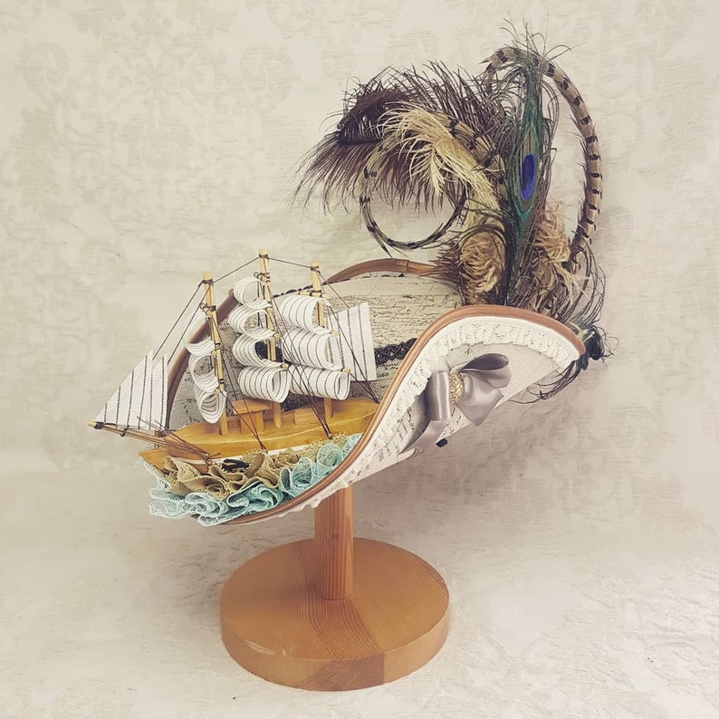 K&ouml;nnte beinhalten: Dekorativer Hut mit einem Miniatur-Holzsegelschiff mit wei&szlig;en Segeln und einer hellblau-beigen R&uuml;schenbasis. Der Hut ist mit Federn, einer Schleife und Spitzenbesatz verziert und wird auf einem Holzst&auml;nder pr&auml;sentiert.