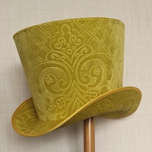 Puede incluir: Un sombrero de copa alto de terciopelo verde con un diseño estampado. El sombrero tiene un ala ancha y una corona alta.