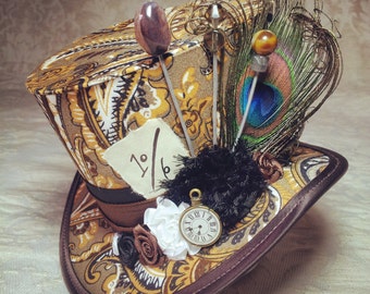 Puede incluir: Un sombrero de copa marrón y dorado con un patrón de paisley y una esfera de reloj en el lateral. El sombrero está decorado con una pluma de pavo real, una boa de plumas negras y una pequeña esfera de reloj.