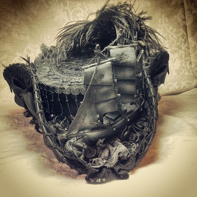May include: A black, ornate, steampunk-style mask featuring a miniature ship with sails. The mask is embellished with feathers, lace, and decorative elements. The design incorporates a mix of textures and intricate details, creating a dramatic and artistic accessory.
