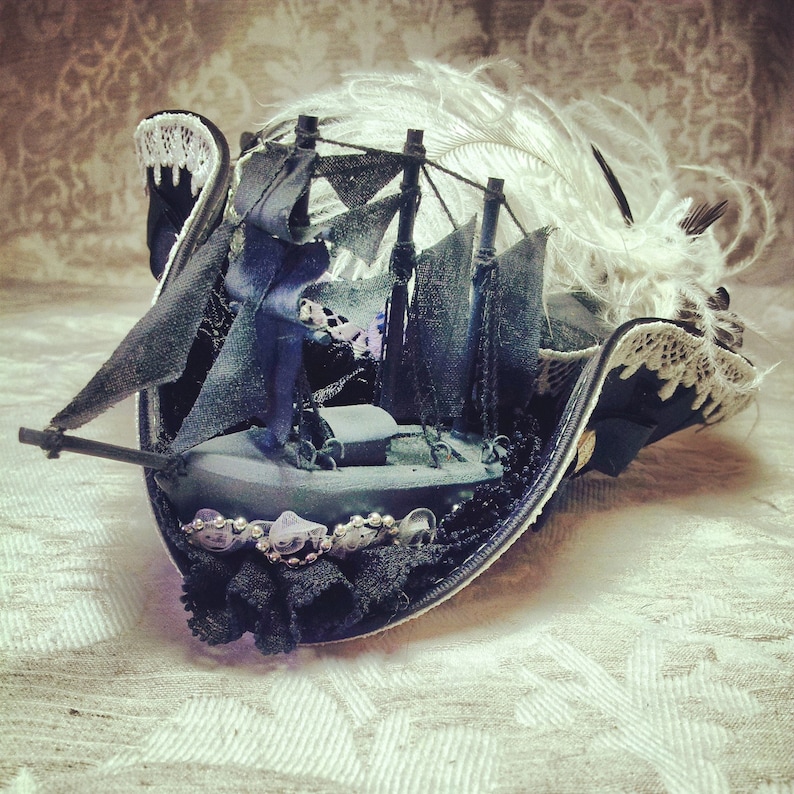 May include: A black pirate hat with a miniature ship scene. The hat features a black brim with white lace trim, a ship with black sails, and white feathers. The hat is decorated with beads and small embellishments.