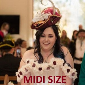 May include: A bride wearing a white wedding dress with a lace bodice and floral embellishments. She is wearing a unique hat shaped like a ship with red, white, and gold accents. The hat is decorated with feathers and a red bow. The bride is walking down the aisle with a smile on her face. The text "MIDI SIZE" is visible in the image.