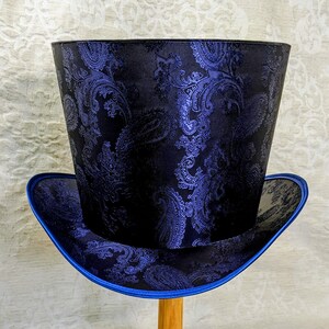 May include: A tall top hat with a black paisley pattern and a blue brim. The hat has a cylindrical crown and a wide, flat brim. The paisley design is a dark blue color.