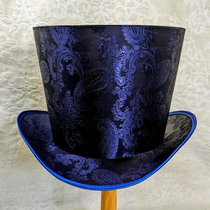 May include: A tall top hat with a black paisley pattern and a blue brim. The hat has a cylindrical crown and a wide, flat brim. The paisley design is a dark blue color.