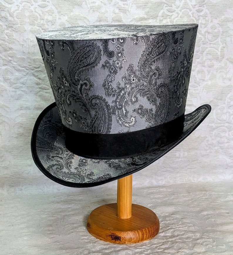 May include: A gray top hat with a paisley pattern and a black band. The hat is on a wooden stand.