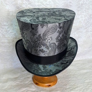 May include: A black and silver top hat with a paisley pattern. The hat is sitting on a wooden stand.
