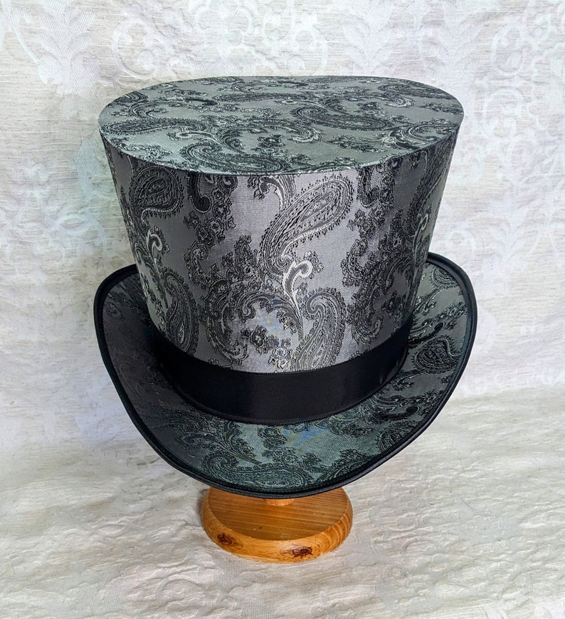 May include: A black and silver top hat with a paisley pattern. The hat is sitting on a wooden stand.