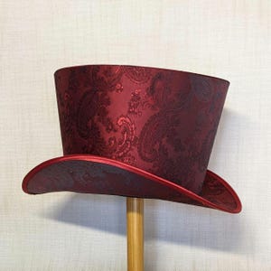 K&ouml;nnte beinhalten: Ein bordeauxfarbener Zylinderhut mit Paisley-Muster. Der Hut hat eine breite Krempe und eine hohe, zylindrische Krone. Der Rand und der Rand der Krone sind mit einem roten Satinband verziert. Der Hut wird auf einem Holzst&auml;nder ausgestellt.