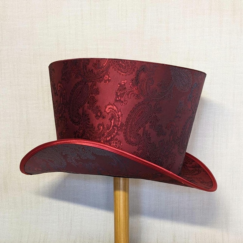 K&ouml;nnte beinhalten: Ein bordeauxfarbener Zylinderhut mit Paisley-Muster. Der Hut hat eine breite Krempe und eine hohe, zylindrische Krone. Der Rand und der Rand der Krone sind mit einem roten Satinband verziert. Der Hut wird auf einem Holzst&auml;nder ausgestellt.